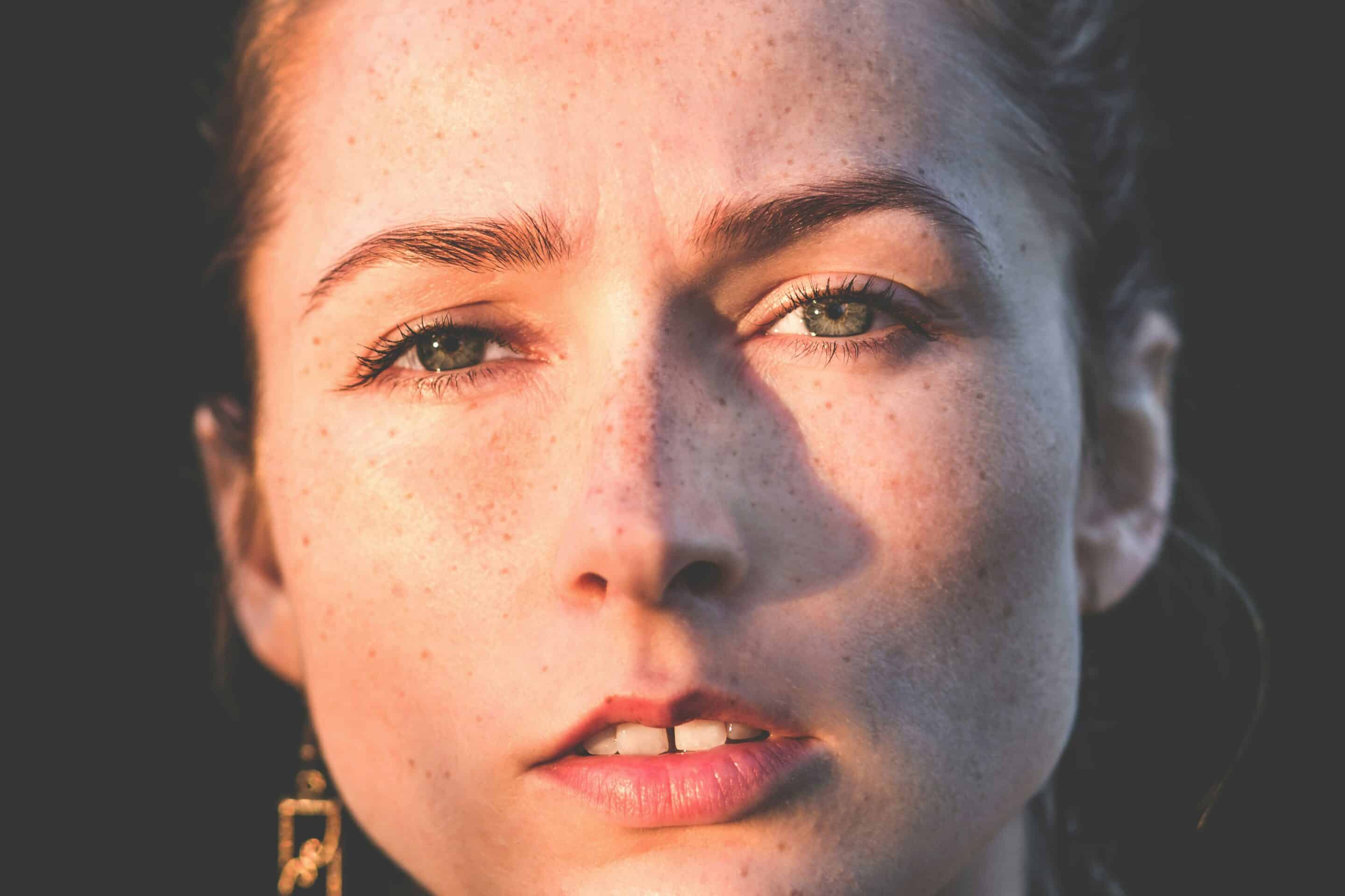 A close-up of a woman’s face with freckles, green eyes, and sunlight casting a shadow across her nose and cheeks.