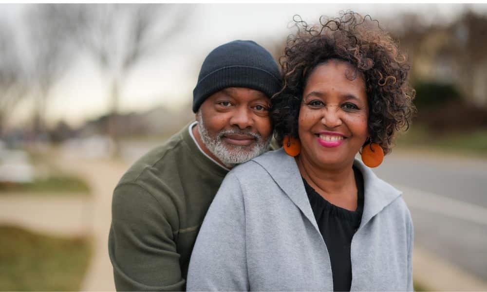 Smiling couple outdoors, with a man in a black beanie hugging a woman in a grey jacket from behind, used to illustrate a grey tooth in cosmetic dentistry