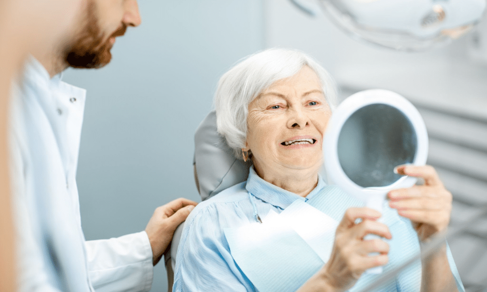Older woman in a dental chair to fix smile ageing smiling while holding a hand mirror, with a dentist beside her in a bright clinic.